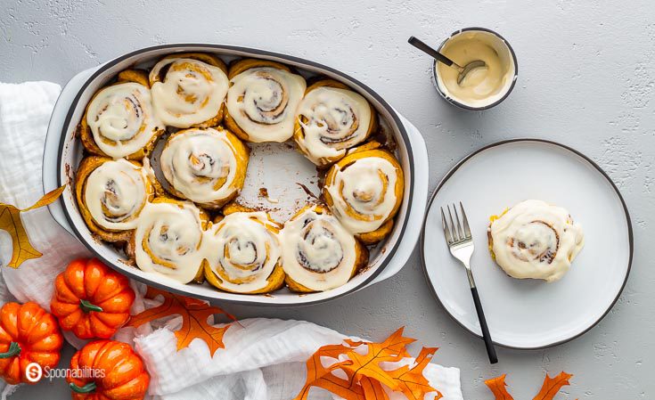 Overhead photo with a baking dish with ten cinnamon rolls and one plate with one pumpkin cinnamon roll. A small container with the maple cream cheese icing. Recipe at Spoonabilities.com