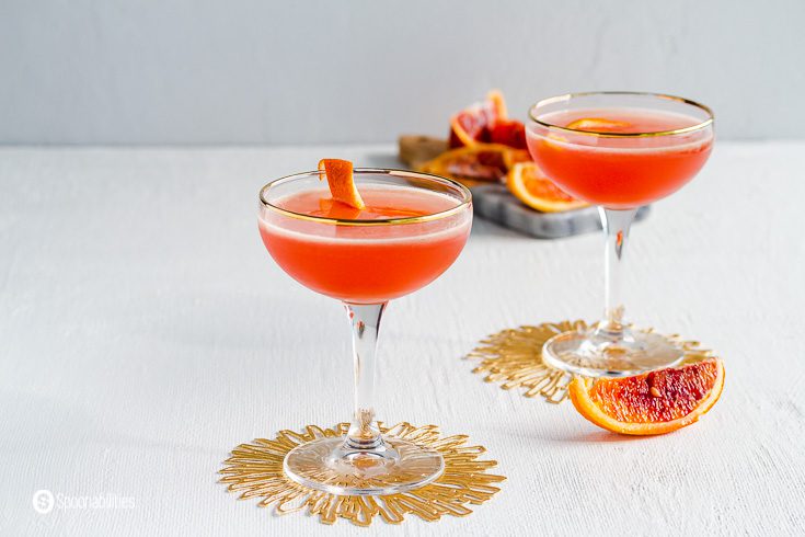 Close up of two couple glasses on top of a golden coasters. A wedge of fresh blood orange next to the glasses. In the background a cutting board with more blood orange pieces. Recipe at Spoonabilities.com
