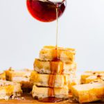 A hands pouring maple drizzle on a tower of cheesecake bars and the drizzle is dripping down looking decadent. Recipe at spoonabilities.com