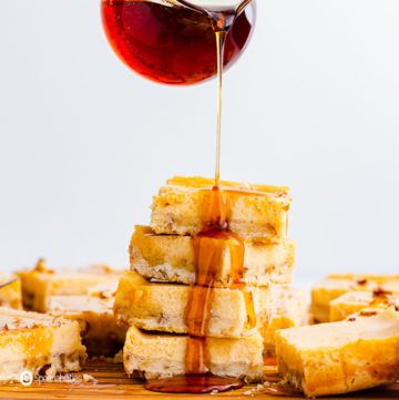 A hands pouring maple drizzle on a tower of cheesecake bars and the drizzle is dripping down looking decadent. Recipe at spoonabilities.com