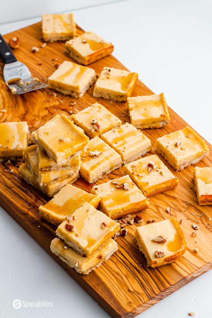 A large olive oil wooden board with 20 plus cheesecake bars with maple drizzle and chopped pecans. Recipe at Spoonabilities.com