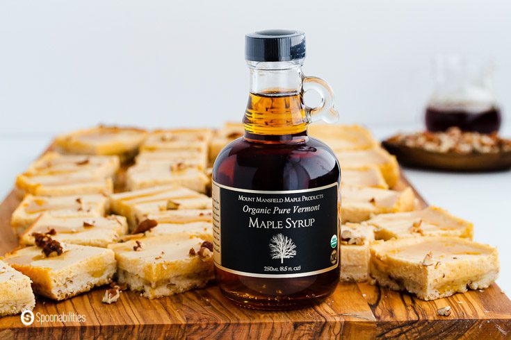 A bottle of Vermont Maple Syrup on top of an wooden board. The wooden board has a tons of Maple Cheesecake bars. Recipe at Spoonabilities.com