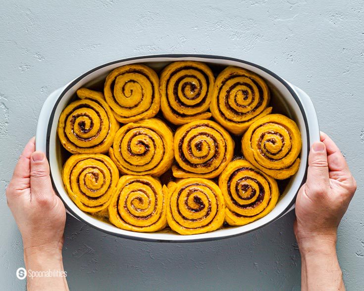 Two hands hold a white oval baking dish with Pumpkin Cinnamon Rolls before adding the maple cream cheese icing. Recipe at Spoonabilities.com