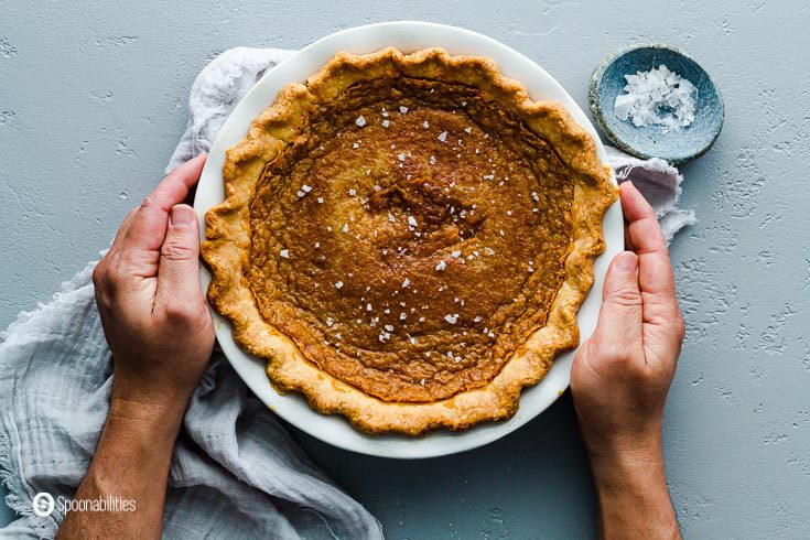 Two hands holding the pie dish with a chess pie inspired recipe. Salted Maple Pie with flaky sea salt. Recipe at Spoonabilities.com