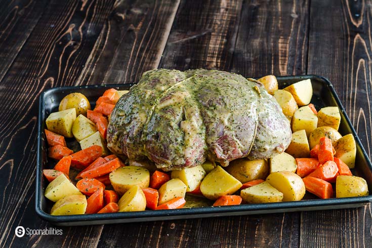 A baking tray with a leg of lamb on top of a bed of Yukon potatoes and carrots ready to go to be roasting on the oven. Recipe at Spoonabilities.com