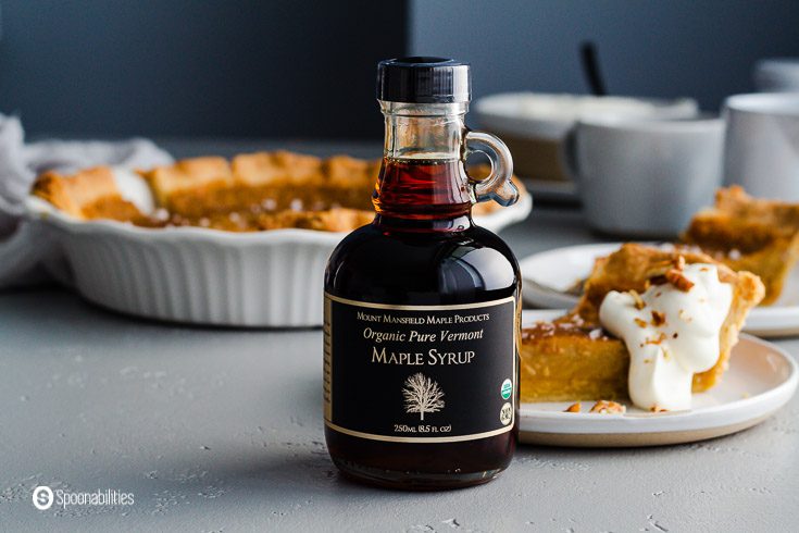 A small bottle of Maple Syrup - Grade B Dark Robust and in the background a pie dish and on the right side two small plates with a piece of the sister pie's cookbook. Recipe at Spoonabilities.com