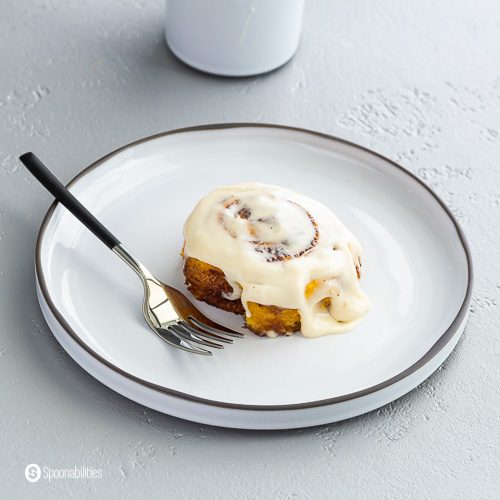 A white round plate with a pumpkin cinnamon roll glazed with maple cream cheese icing with a fork on the plate ready to try the first piece. Recipe at Spoonabilities.com