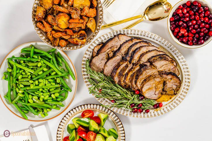 Thanksgiving table landscape with five plates as follow: an oval plate with roasted leg of lamb, tomato & cucumber salad, green beans, roasted vegetables, and a bowl with fresh cranberry. Recipe at Spoonabilities.com