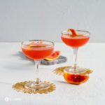 Two Coupe glasses with Solerno Cocktail . The two glasses on top of a golden coasters.