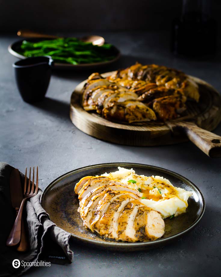 Plate with sliced brown butter roasted turkey breast with mashed potatoes. in the background a wooden board with the rest of the turkey. Recipe at Spoonabilities.com