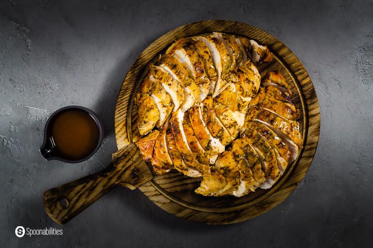 Photo with a wooden serving board with sliced roasted turkey breast and on the left bottom side a vessel with gravy. Recipe at Spoonabilities.com