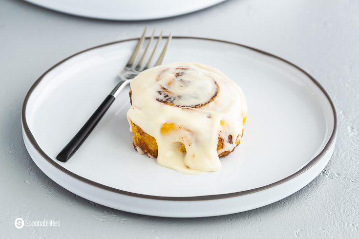 One pumpkin cinnamon roll on a round white plate with maple cream cheese glaze. A fork on the left side of the plate. Recipe at spoonabilities.com