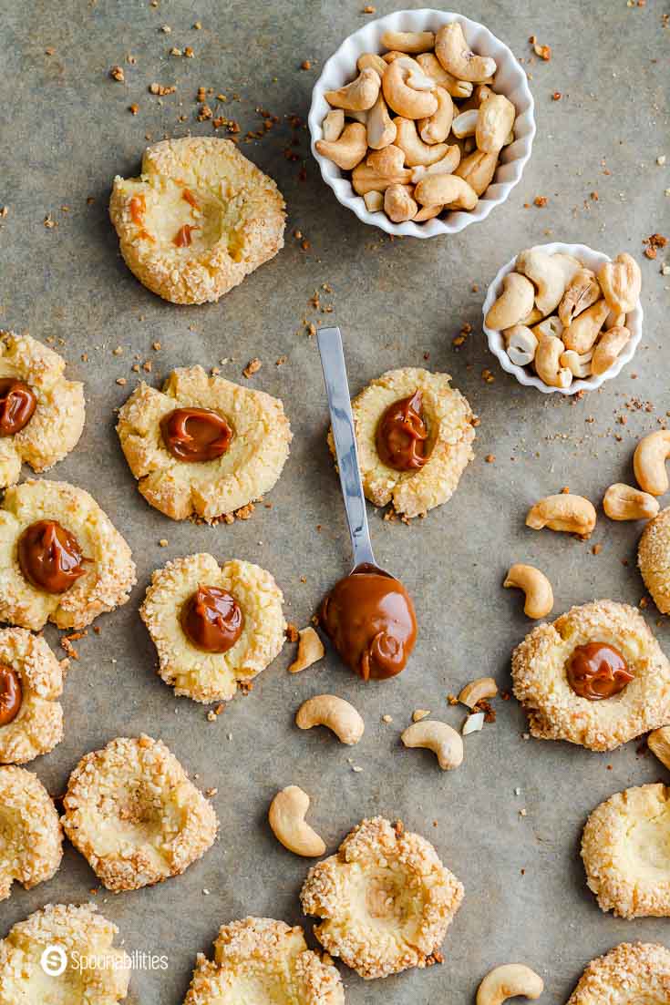 Cashew thumbprint cookies on a baking tray. Some cookies with a spoonful of cajeta sauce and others without the filling. A Spoon with caramel sauce in the center of the photo. Recipe at Spoonabilities.com