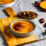 A grey ramekin on top of a wooden board and garnished around some pilled chestnuts in a small wooden pinch bowl. The ramekin has Chestnut Crème Brûlée. Recipe at Spoonabilities.com