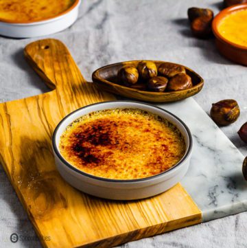 A grey ramekin on top of a wooden board and garnished around some pilled chestnuts in a small wooden pinch bowl. The ramekin has Chestnut Crème Brûlée. Recipe at Spoonabilities.com