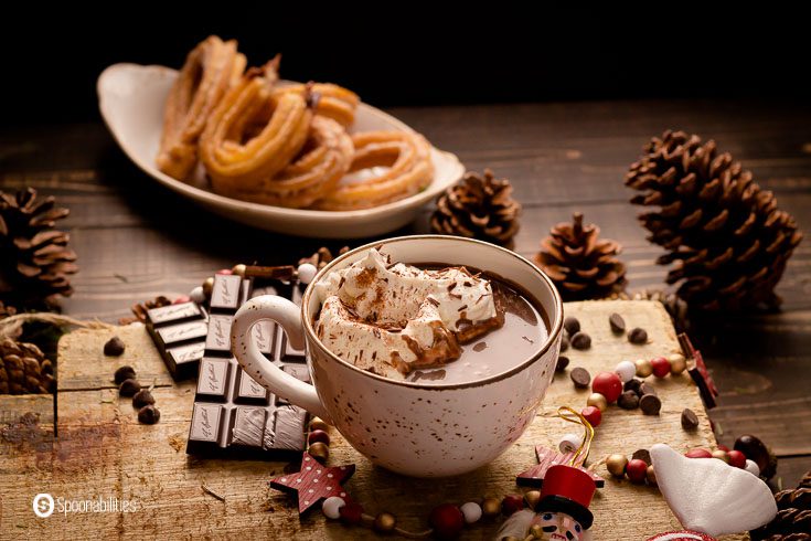 A cozy photo with a Christmas décor with a mug with spiced Spanish hot chocolate with a dollop of whipped cream. Recipe at Spoonabilities.com