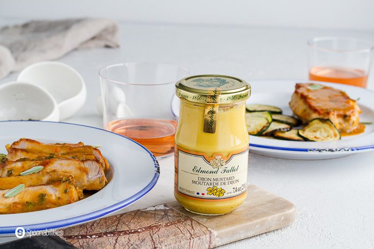 a jar of Edmond Fallot Dijon Mustard between two white plates with pan-seared pork chops with apricot Dijon mustard sauce and zucchinis. Available at our shop at Spoonabilities.com