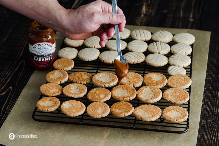 Cookies in a cooling rack and a hand with a spoon with goat's milk caramel sauce is filing the cookies with the sauce. Recipe at Spoonabilities.com