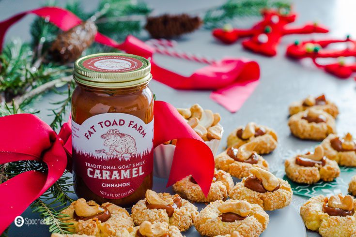 A jar of Cinnamon Goat's milk caramel sauce (Cajeta) with some thumbprint cookies around the jar filled with caramel sauce and a whole cashew on top. The jar is with a red bow. Recipe at Spoonabilities.com