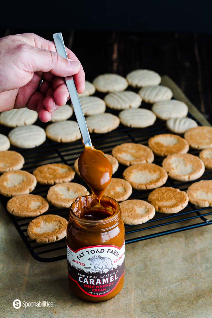 A open jar of Caramel sauce and a hand with a spoon with caramel sauce. in the background the shortbread cookies in a cooling rack ready to be filled. Recipe at Spoonabilities.com