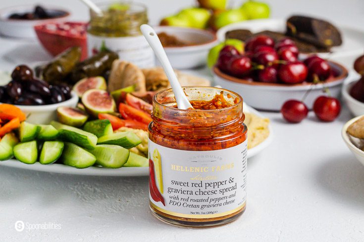 A open jar of sweet red pepper and graviera cheese spread from Hellenic Farms. The jar has a white spoon inside. In the background a large plate with some mezze dishes. Product available at Spoonabilities.com