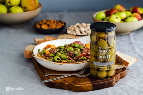 A jar of Green green olives on a wooden tray and in the background a plate with marinated olives, a bowl of apples, pears, nuts, and bread. Recipe at Spoonabilities.com