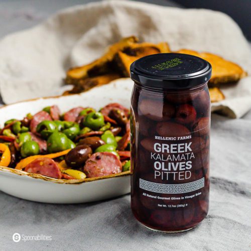 A jar of pitted Kalamata olives and in the back you will see a small plate with warm marinated olives with salami. Product available at Spoonabilities.com