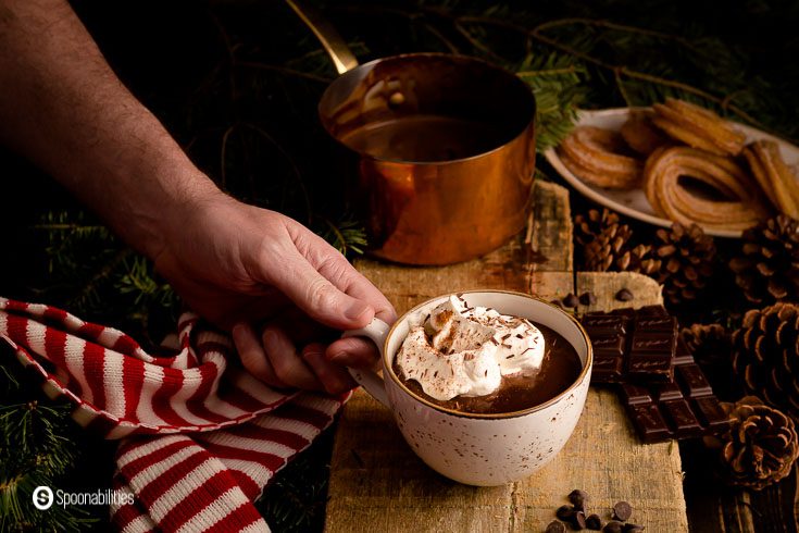 A hand holding a cup of Spiced Spanish hot chocolate. There is a holiday cozy décor in the photo. Recipe at Spoonabilities.com
