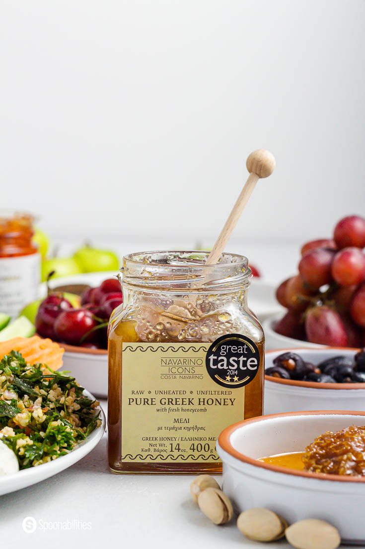 An open jar of pure Greek Honey with a honeycomb inside. The jar is next to some small dishes with Greek appetizers. Recipe at spoonabilities.com
