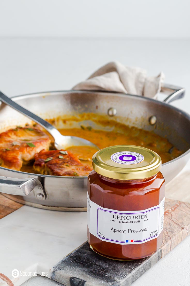 A close up photo of L’Epicurien Apricot Preserves jar. The jar is on top of a marble board and in the background a sauté pan with two pork chops and one spoon. Available at our shop at spoonabilities.com