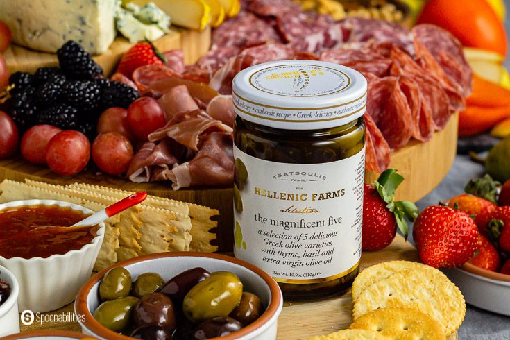 Jar of The Magnificent Five Greek olives on a wooden charcuterie board with cured meats.