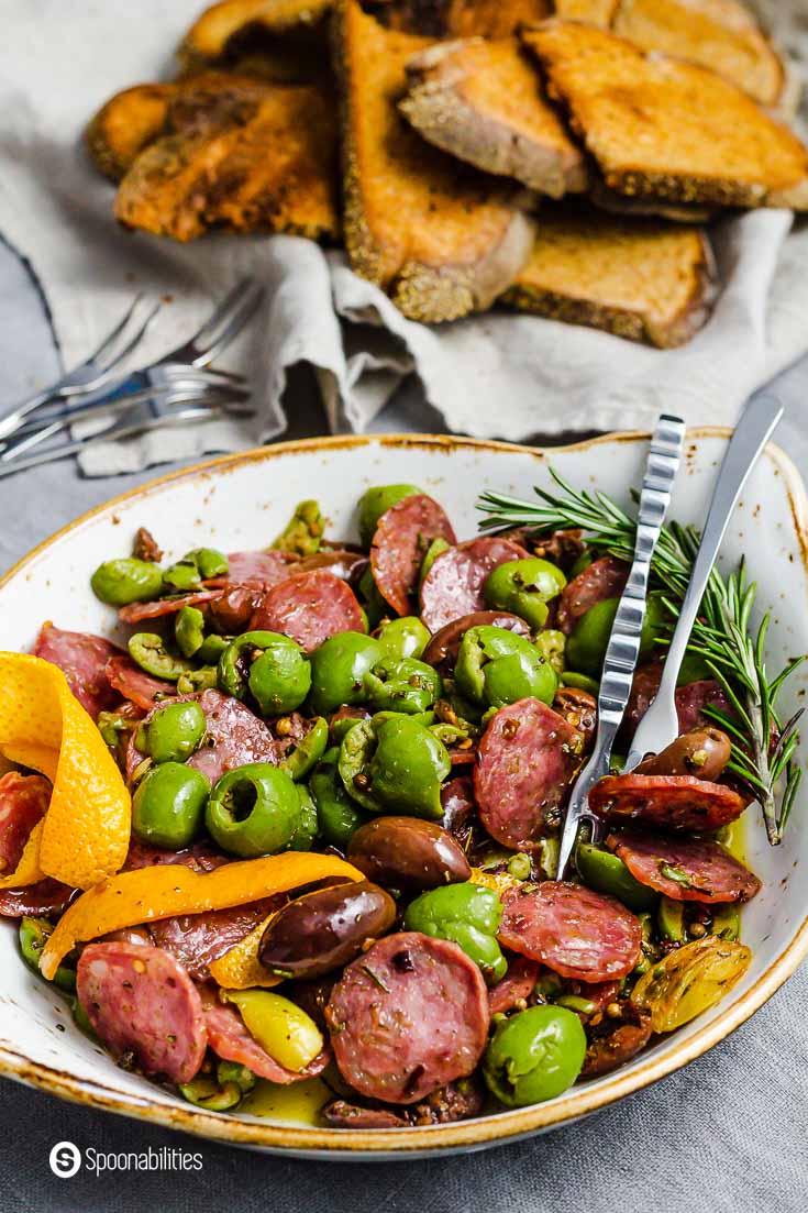 Close up photo of a plate. The plate has warm olives marinated in olive oil, spices, herbs, and salami. Recipe at Spoonabilities.com