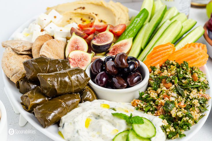 Mezze platter of a white plate with Greek appetizers like dolma, tabbouleh, olives, hummus, figs, cucumbers, feta cheese, and pita bread. Recipe at Spoonabilities.com