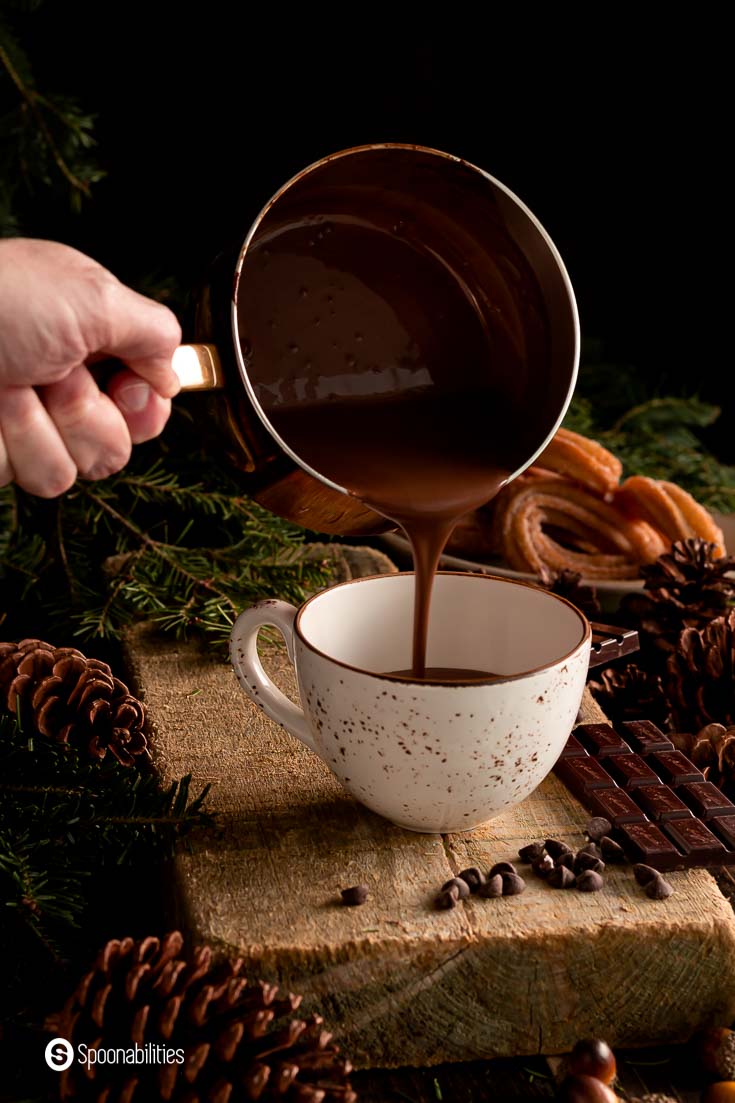 Pouring Spanish hot chocolate into a mug. The mug is on top of a wooden block and around the mug is decorate with a cozy holiday décor. Recipe a Spoonabilities.com
