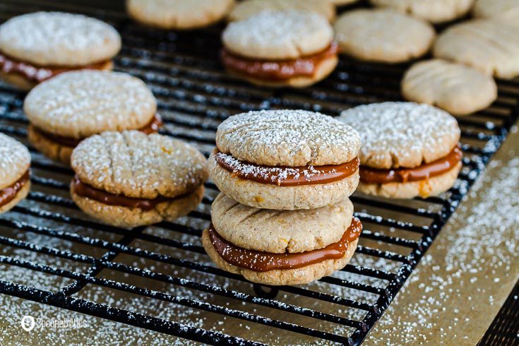 Black wire cooling rack with shortbread cookies filled with Goat's milk caramel sauce flavored with Irish Whiskey cream. The main cookies are in the center of the photo with two sandwiches cookies on top of each other. Recipe at spoonabilities.com