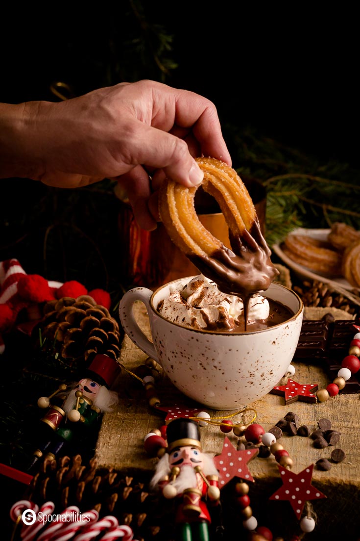 A cozy mug with hot chocolate and a hand dipping a churros into the Spiced Spanish Hot Chocolate. Recipe at Spoonabilities.com