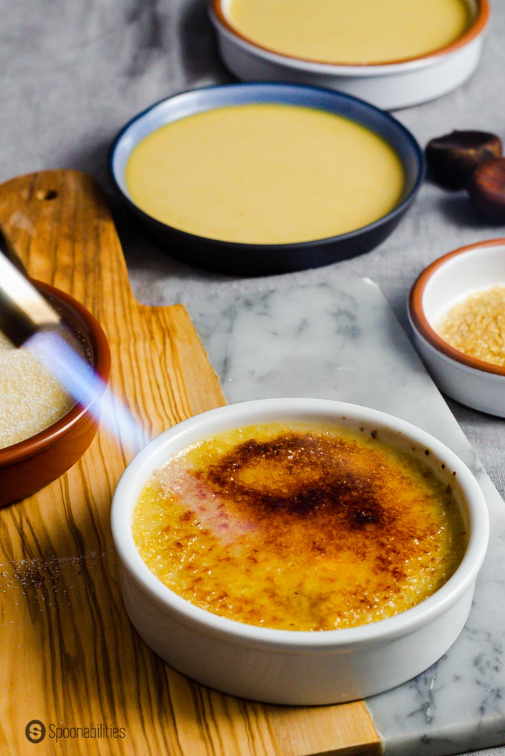 A white ramekin with Crème brûlée with sugar on top and a hand torching the brûlée. You can see the caramelized sugar layer on top of the custard. Recipe at Spoonabilities.com