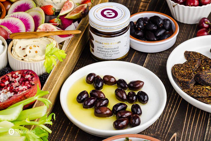 A jar of Kalamata olives marinated in anise and other Mediterranean spices. In front of the jar a small plate with the olives and olive oil from the marinated. Product at Spoonabilities.com