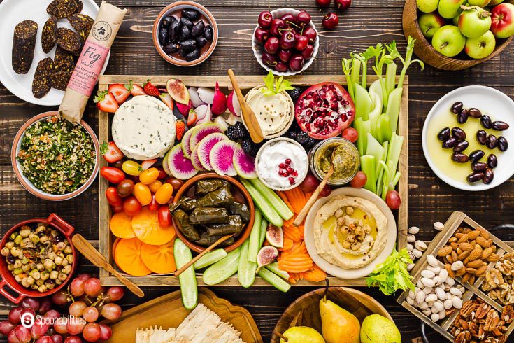 A grazing table with a lot of vegan appetizers in a large wooden tray and small plates with fruits, salads, and nuts. Recipe at Spoonabilities.com