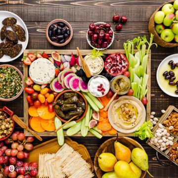 A grazing board with a lot of vegan appetizers in a large wooden tray and small plates with fruits, salads, and nuts. Recipe at Spoonabilities.com