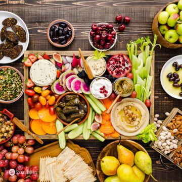 A grazing board with a lot of vegan appetizers in a large wooden tray and small plates with fruits, salads, and nuts. Recipe at Spoonabilities.com