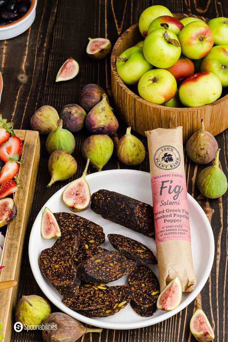 Round white plate with slices of vegan fig salami and next a whole vegan salami with the label in the original packaging. Behind the plate fresh figs. Product available at Spoonabilities.com