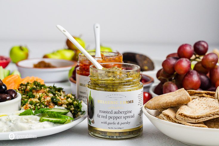 A open jar of roasted Aubergine and red pepper spread from Hellenic Farms. The jar has a white spoon inside. Around the jar are plates with some mezze dishes. Product available at Spoonabilities.com