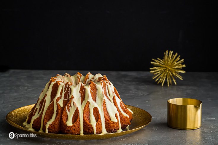 A photo with a Eggnog Bundt Cake on a plate with golden rim and on the right side a golden pouring cup with the glaze with brandy flavoring. Recipe at Spoonabilities.com
