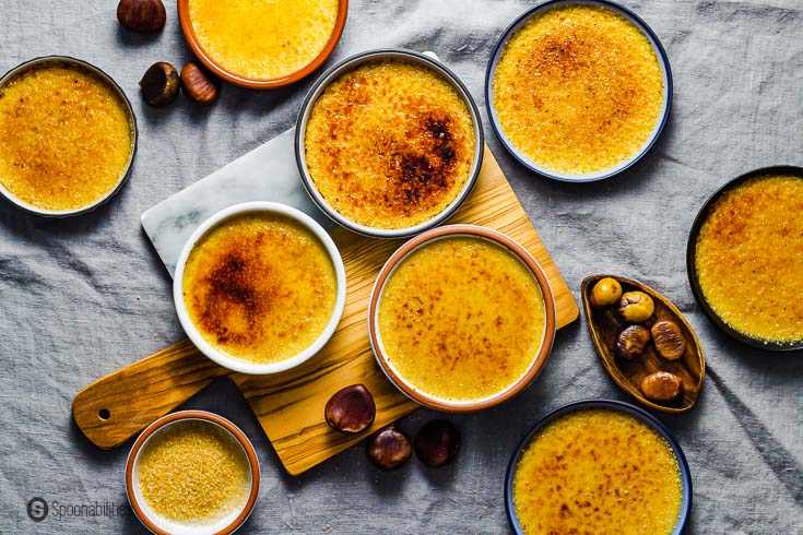 Eight ramekins on a table with a grey tablecloth with Chestnut Crème Brûlée with burnt Demerara sugar. Three of the ramekins are on top of a wooden board and there are some fresh roasted chestnuts around the table. Recipe at spoonabilities.com
