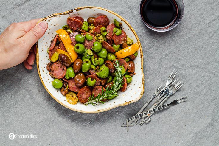 A hand holding a plate with the easy appetizer recipe Warm marinated olives. Next to the plate a appetizer forks and a glass of red wine. Recipe at Spoonabilities.com