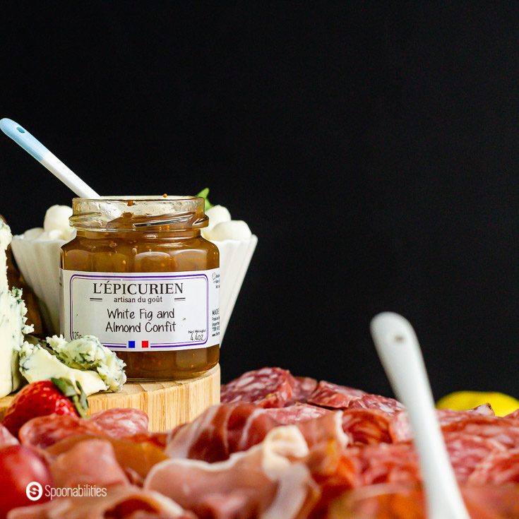 A jar of white fig and almonds confit next to blue cheese surrounded by cured meats as part of a charcuterie board recipe.