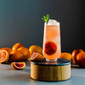 Beautiful tall glass with a blood orange cocktail topped with ginger beer. The glass is on top of a wood/marble pedestal, and in the background, some fresh blood oranges. Recipe for this ginger beer cocktail at Spoonabilities.com