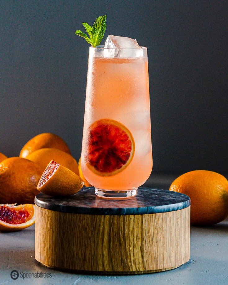 A beautiful tall cocktail glass in a wooden pedestal and in the background fresh blood oranges. Recipe at Spoonabilities.com