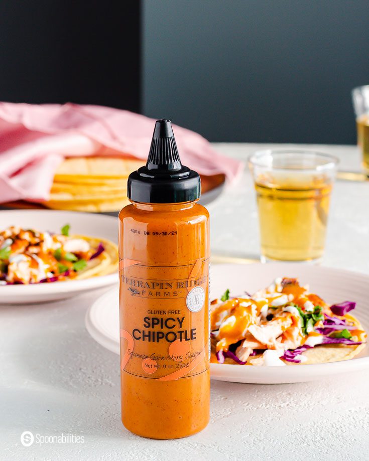 A Squeeze bottle of Spicy Chipotle sauce from Terrapin Ridge, the bottle is in front of a pink plate with a taco. This sauce is available at Spoonabilities.com